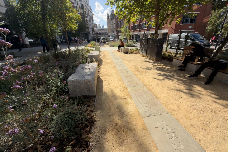 A photograph of the Hicks Hall memorial on a sunny day. It shows the 11 metre long path of 19 reclaimed yorkstone paving slabs sandblasted with the story of Hicks Hall. These sit on a wider sandy gravel path within a pocket park, with stone seating and rainwater beds planted with trees and wildflowers. In the background you can see St. John Street and Smithfield market.