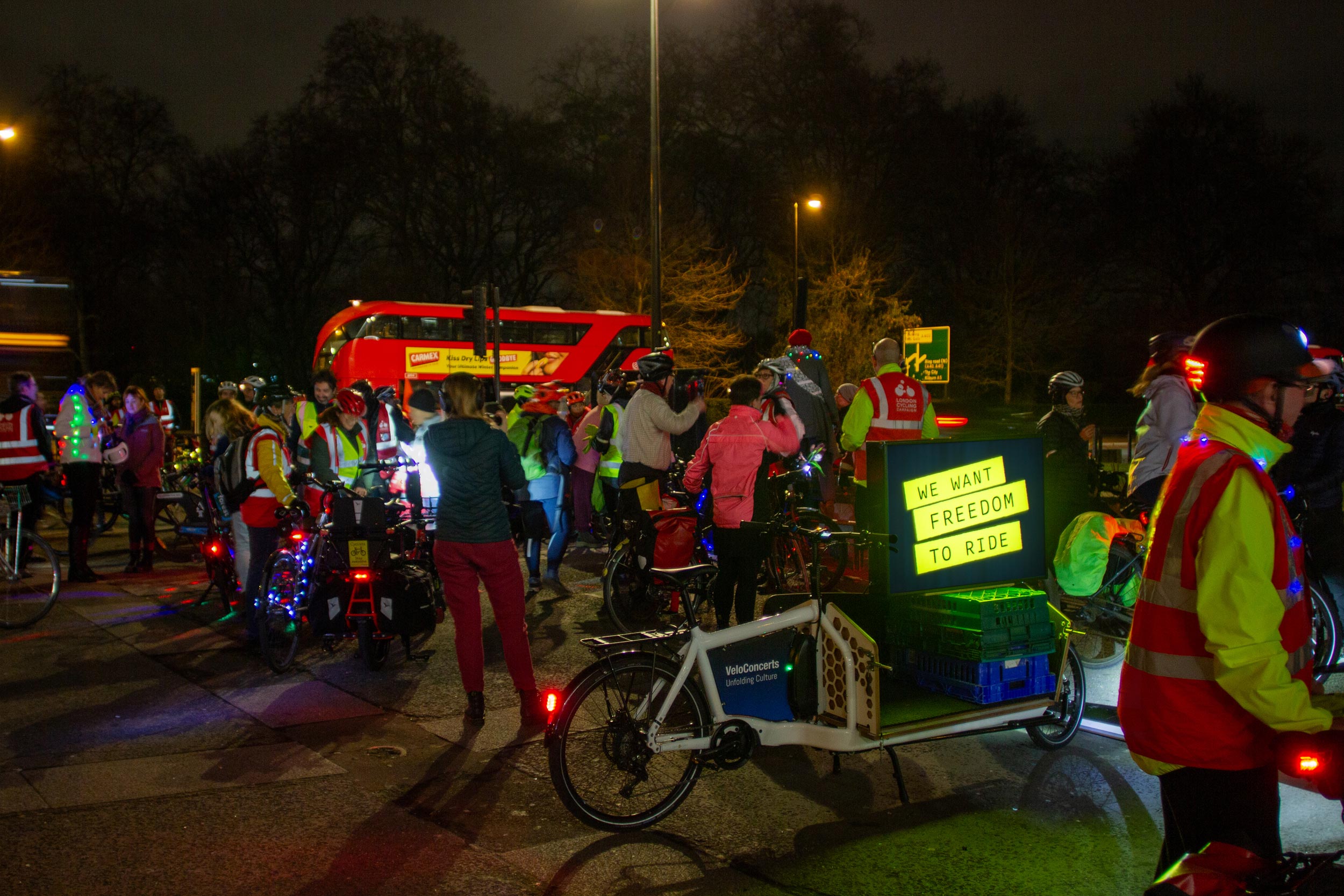 A group of cyclists gathered at night at Marble Arch in London, with a cargobike with a lightbox mounted on it, on which there's a logo which reads 'We want freedom to ride'.