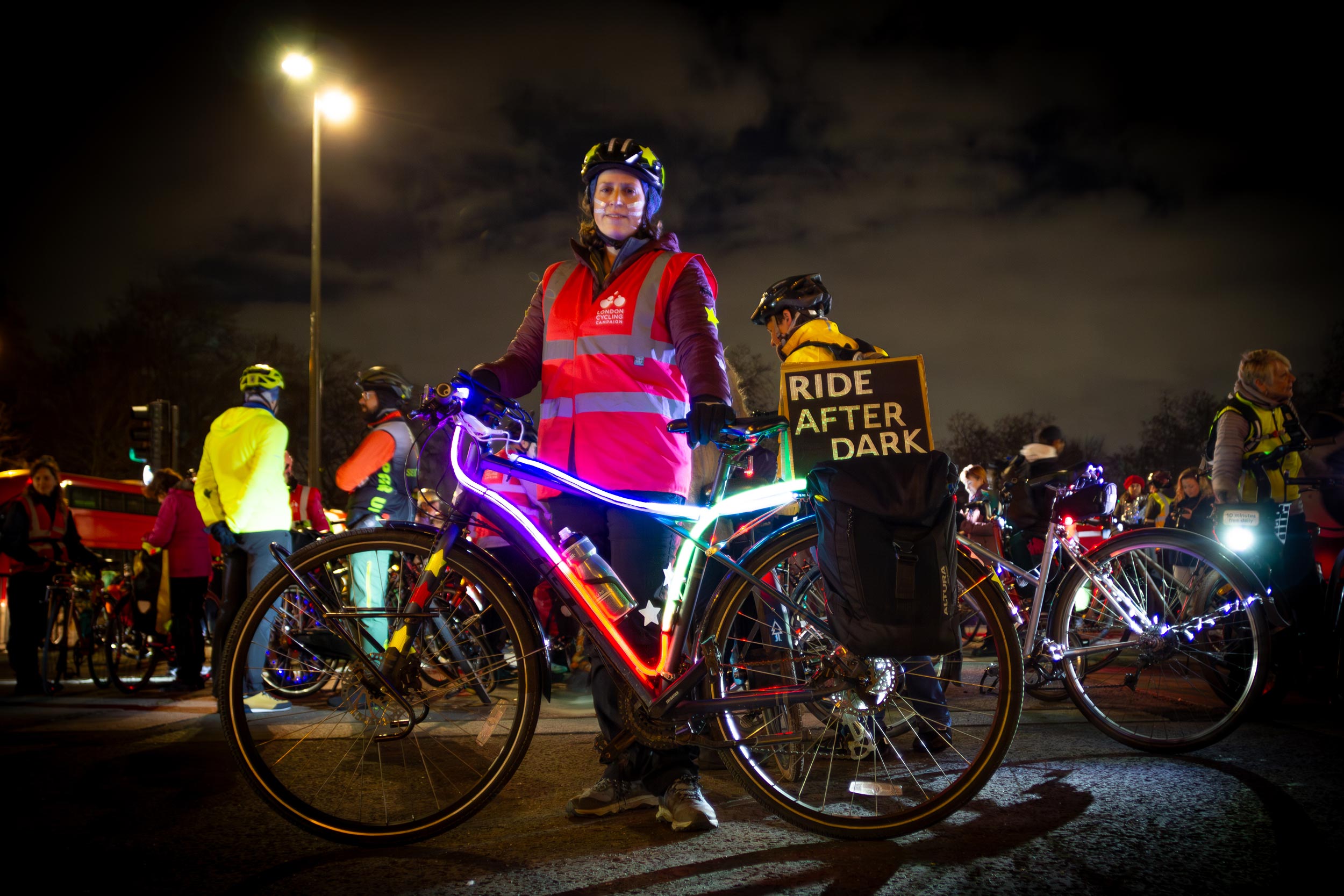 A woman standing behind her bike, which is lit up with multi coloured neon lights, and a with a 'Ride after dark' handmade sign.