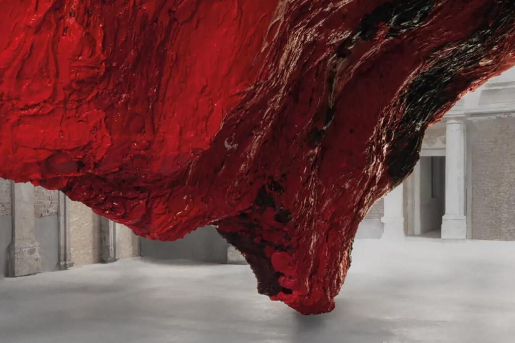 A photograph of a huge red organic sculpture by Anish Kapoor, within a gallery space. Title: 'Mount Moriah at the Gate of the Ghetto'. Anish Kapoor 2022.