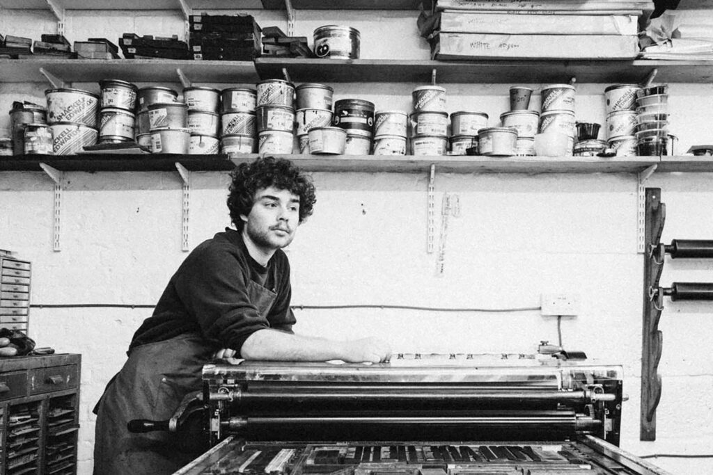 A black and white phogotraph of Theo Hersey leaning on a printing press in his studio.