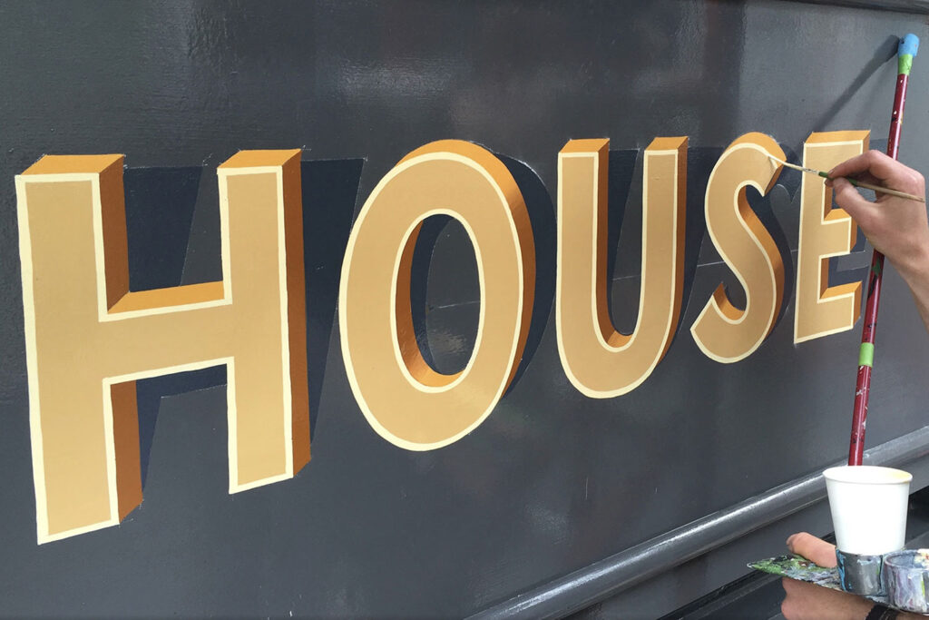 A photograph of the word HOUSE being hand painted in three dimensional lettering on a grey signboard.