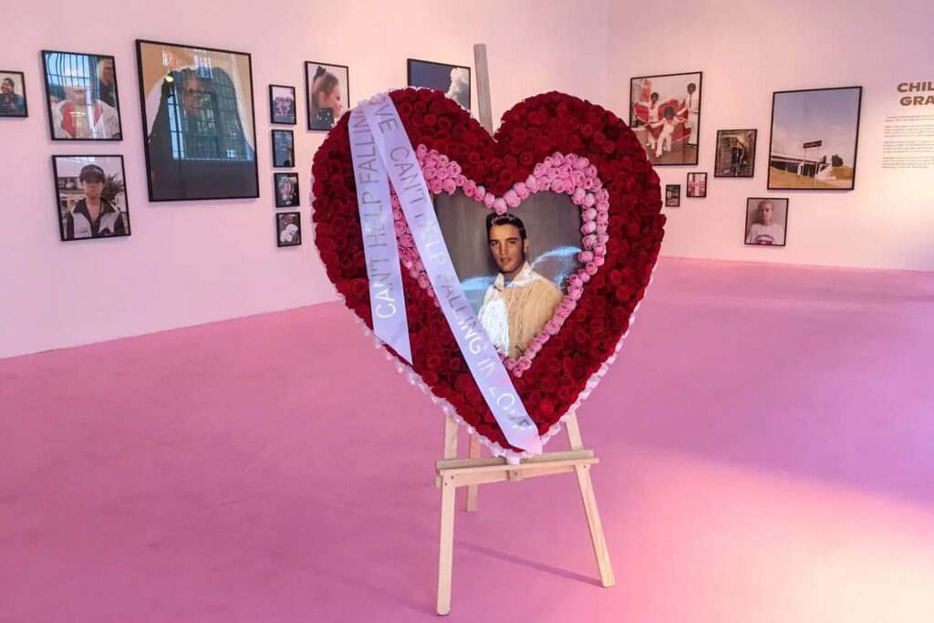 A photograph of a large heart shaped floral wreath in an exhibition space. At the centre of the wreath is a photo of Elvis Presley. Title: 'Graceland exhibition' by Hayley Louisa Brown