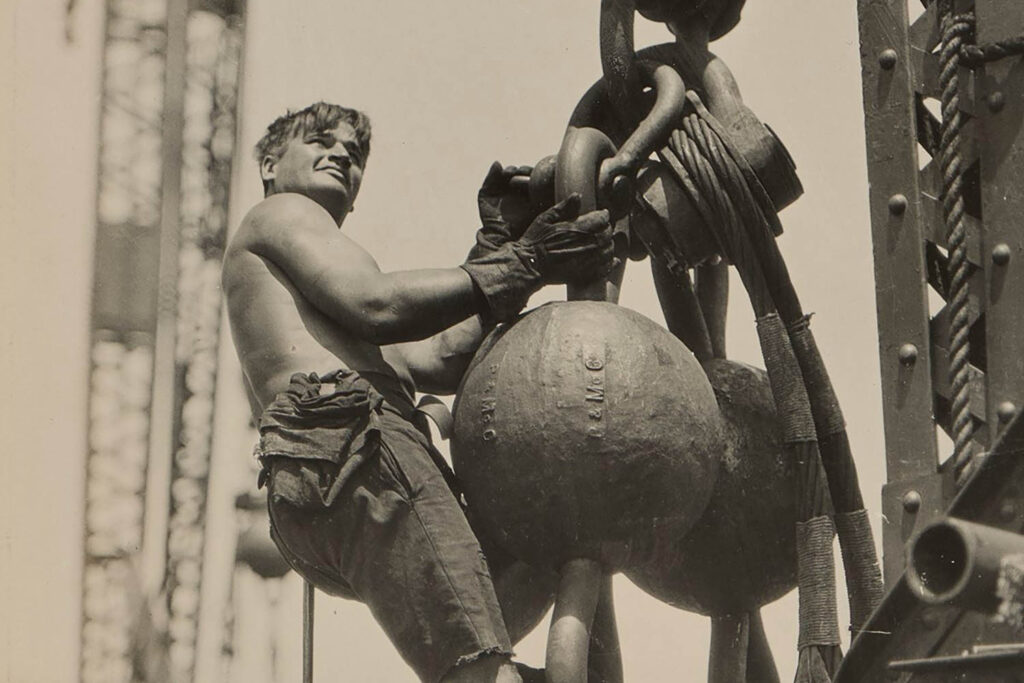 A black and white photograph of a shirtless man standing on the hook of a crane with two large spherical weights. Lewis Hine, Riding the Ball High up on Empire State, c.1930