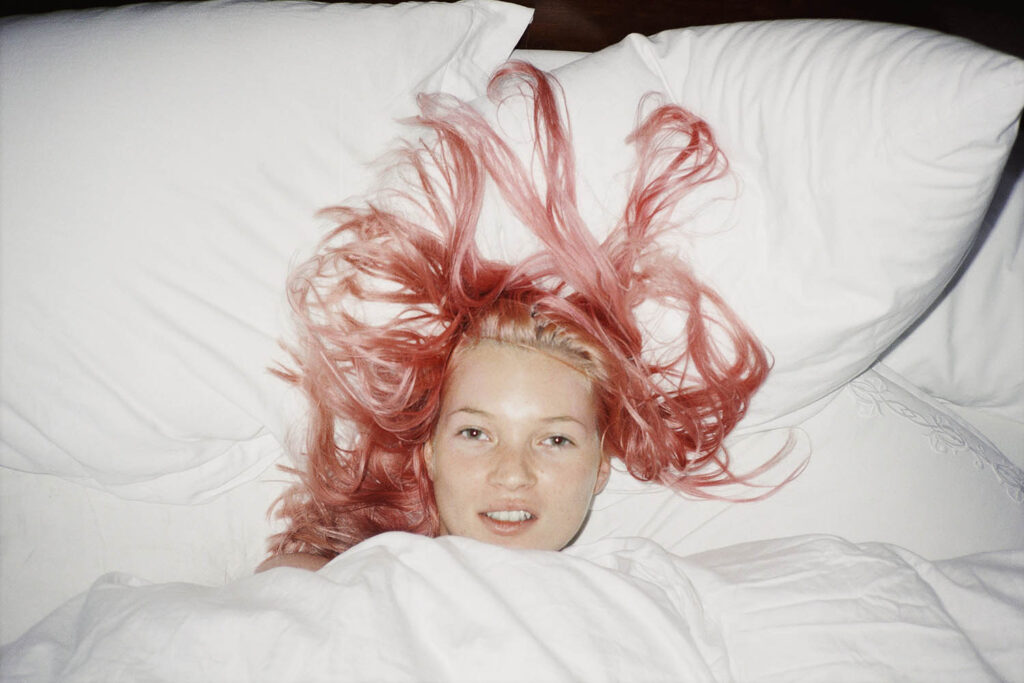 A photograph of Kate Moss in bed, with just her head visible, and her bright pink hair spread out on the pillows. Juergen Teller, Young Pink Kate, London 1998