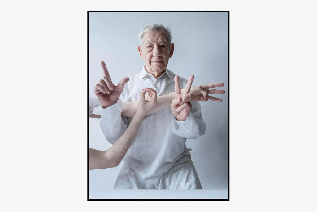 A photograph by Tim Walker of Sir Ian McKellen wearing a white shirt and lace robe, and holding up his hands to make the shapes of an L and a V. The arms of someone else are also visible, making the shapes of an O and an E. Together they spell out LOVE. Ian McKellen, Love, London, 2023.